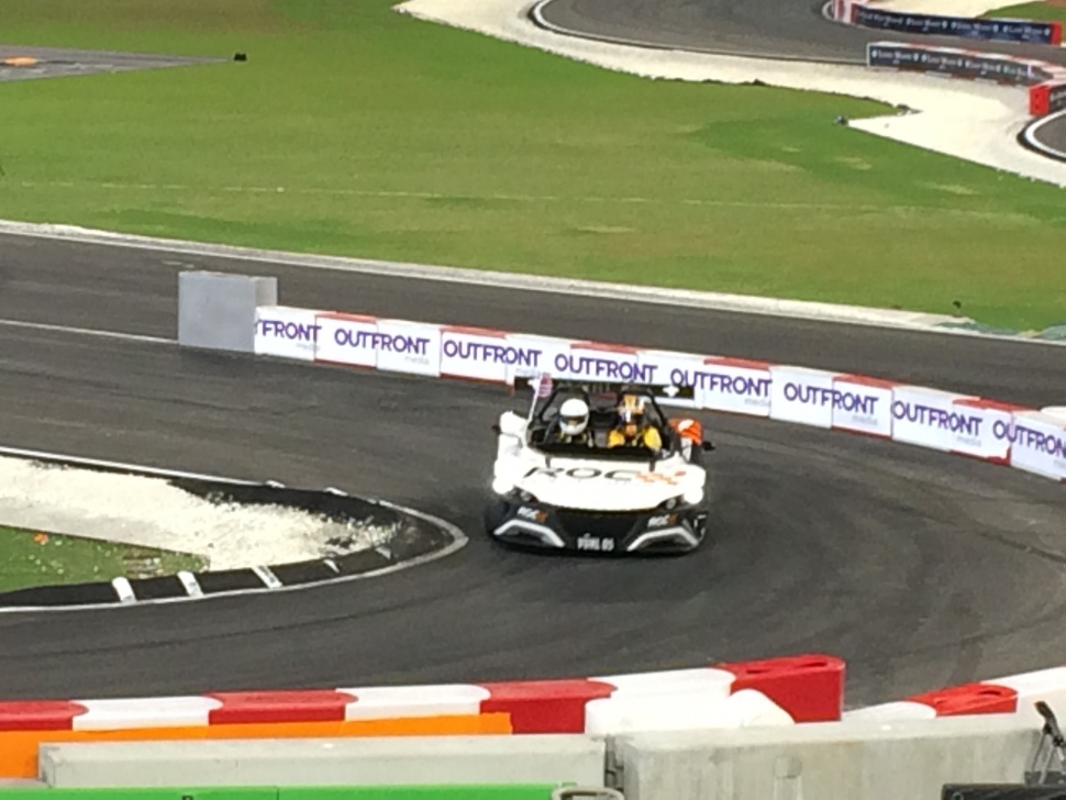 Lightweight open-top roadster with two helmeted drivers on a tight race track bend.