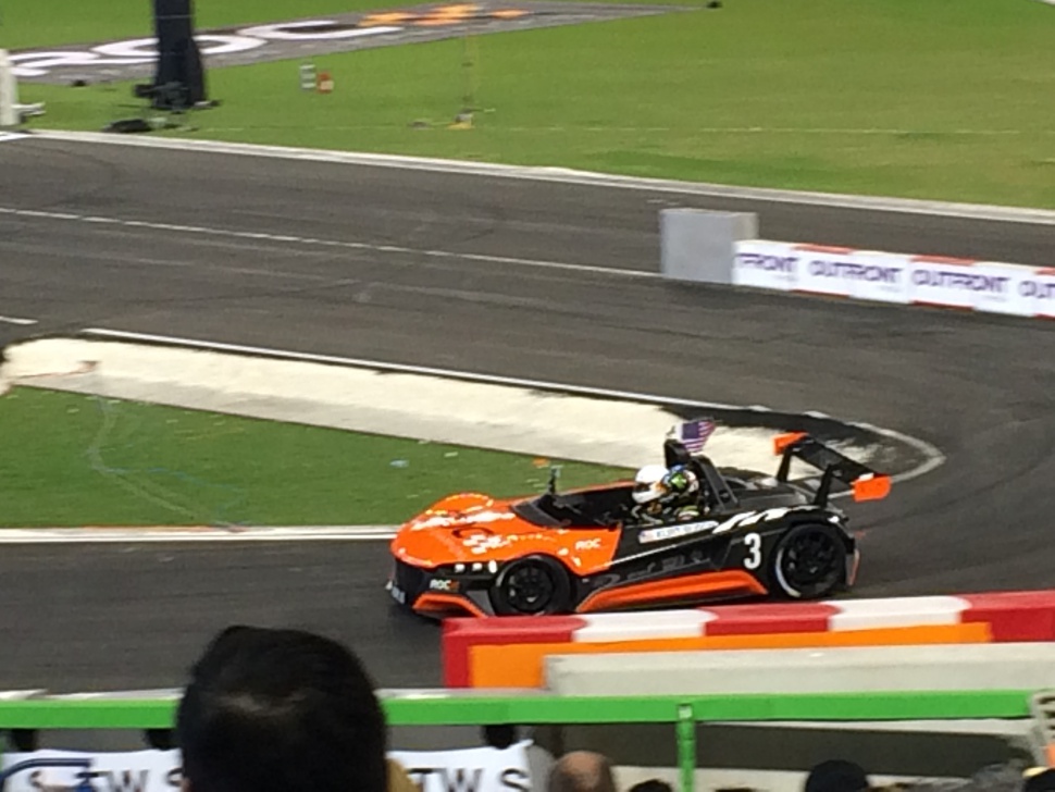 Open-top race car with large rear wing on race track corner.