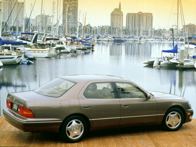 Sedan parked on wooden dock by marina with yachts and waterfront buildings.
