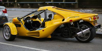 Yellow three-wheeled open cockpit vehicle with exposed rear mechanics and chrome exhausts